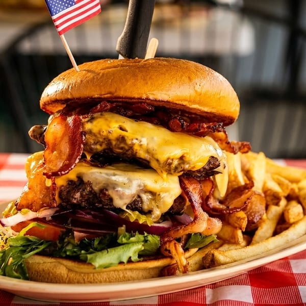 Massive double cheeseburger with bacon and fries, topped with an American flag, on a red checkered tablecloth.
