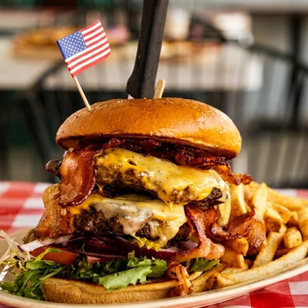 Massive double bacon cheeseburger with fries, topped with an American flag, on a red checkered tablecloth.