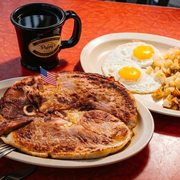 A diner breakfast with a large ham steak, two fried eggs, and crispy hash browns on a red table.